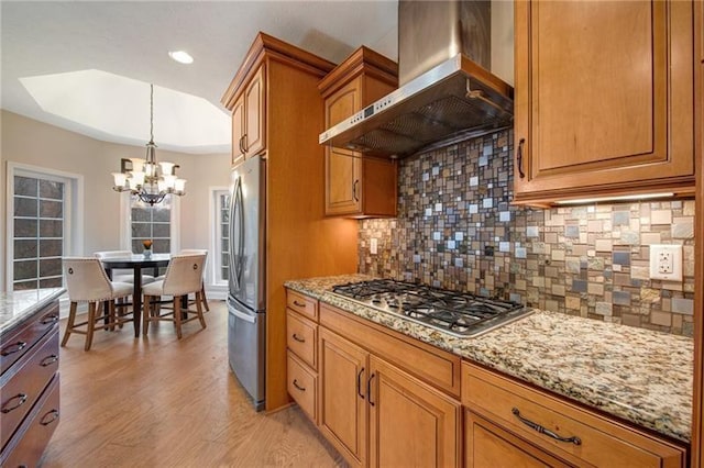 kitchen featuring tasteful backsplash, an inviting chandelier, wall chimney range hood, appliances with stainless steel finishes, and light stone counters