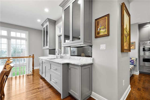 kitchen featuring glass insert cabinets, double oven, a sink, gray cabinetry, and baseboards