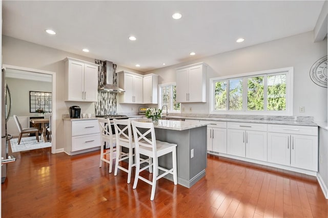 kitchen with white cabinets, a center island, wall chimney exhaust hood, dark wood finished floors, and a breakfast bar area