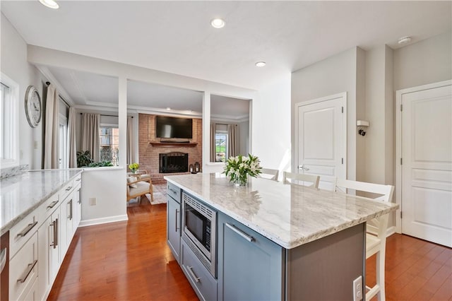 kitchen with stainless steel microwave, light stone countertops, dark wood-type flooring, a brick fireplace, and a kitchen bar