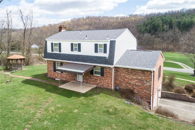 back of house featuring a patio, a view of trees, a chimney, a yard, and a shingled roof