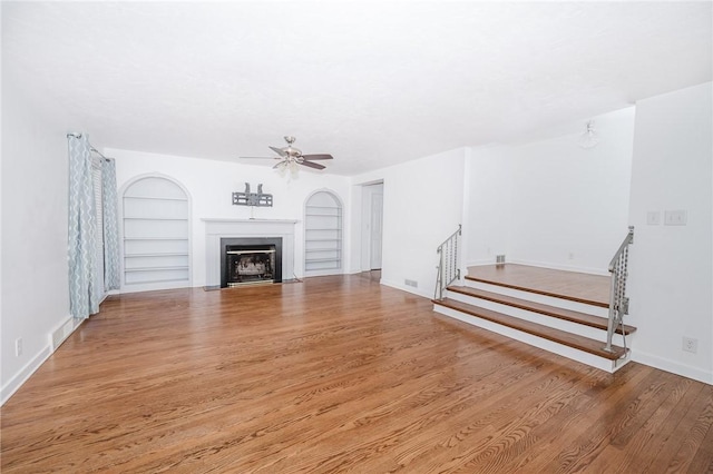 unfurnished living room featuring built in features, a ceiling fan, stairway, wood finished floors, and a fireplace with flush hearth
