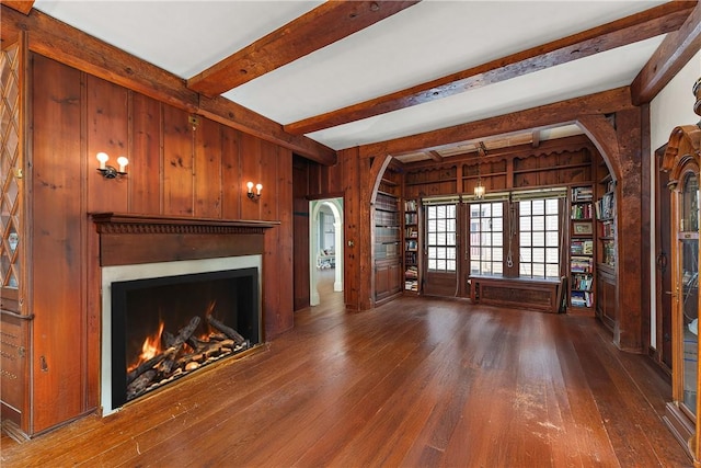 unfurnished living room with arched walkways, a warm lit fireplace, hardwood / wood-style flooring, beamed ceiling, and wood walls