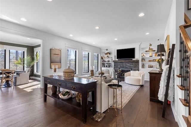 living area with built in features, a stone fireplace, dark wood-style floors, recessed lighting, and stairs