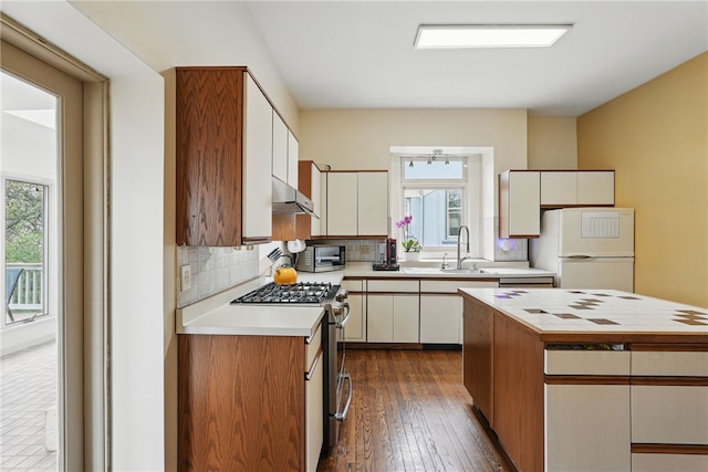 kitchen with tasteful backsplash, light countertops, freestanding refrigerator, under cabinet range hood, and stainless steel gas stove