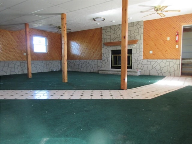 basement featuring wooden walls, a fireplace, a ceiling fan, and carpet