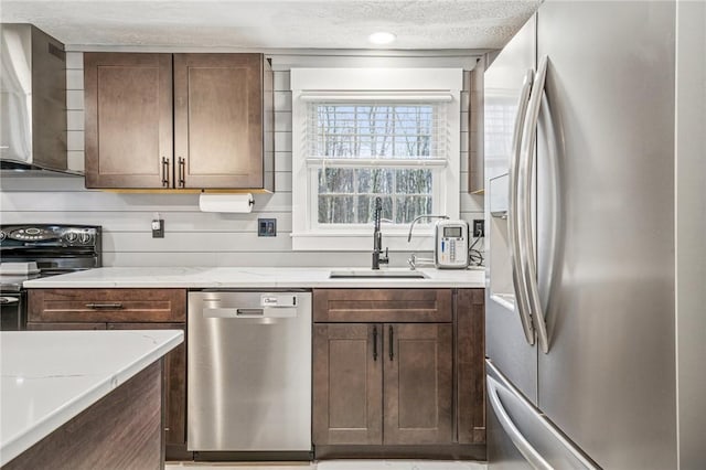 kitchen featuring wall chimney exhaust hood, a textured ceiling, light stone counters, a sink, and stainless steel appliances