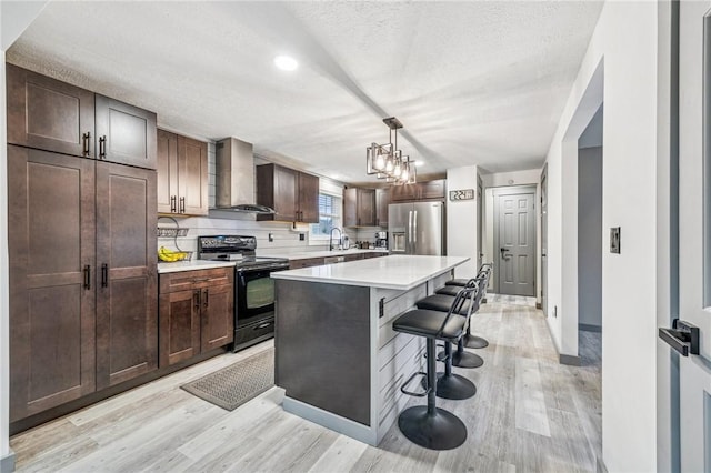 kitchen featuring wall chimney exhaust hood, a kitchen breakfast bar, light wood-style flooring, stainless steel refrigerator with ice dispenser, and black range with electric cooktop