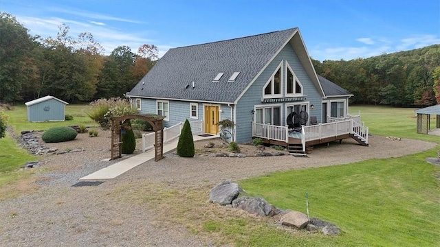 back of property featuring a shingled roof, a wooded view, a lawn, an outbuilding, and a wooden deck