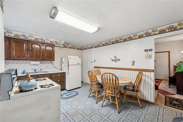 kitchen with dark brown cabinetry, wallpapered walls, white appliances, light countertops, and a sink