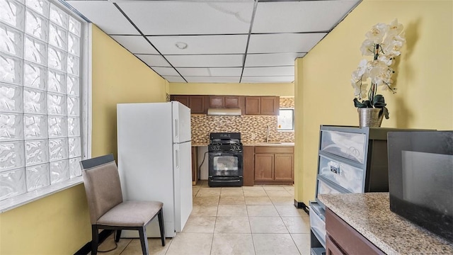 kitchen featuring light tile patterned floors, freestanding refrigerator, under cabinet range hood, brown cabinetry, and black range with gas stovetop