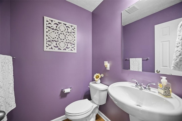 half bath featuring a textured ceiling, visible vents, toilet, a sink, and baseboards