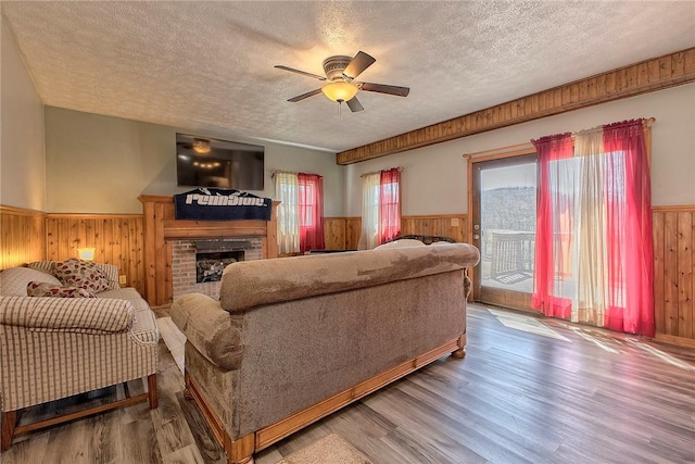 living room featuring wooden walls, a textured ceiling, a wainscoted wall, a brick fireplace, and ceiling fan