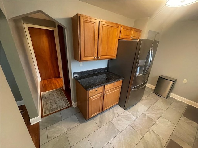 kitchen featuring dark stone counters, brown cabinetry, stainless steel fridge, marble finish floor, and baseboards