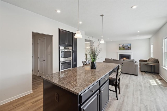 kitchen with a center island, stainless steel double oven, light wood-type flooring, a lit fireplace, and baseboards