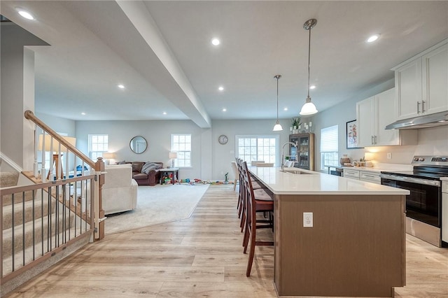 kitchen featuring a sink, under cabinet range hood, light wood-style floors, stainless steel electric range oven, and a center island with sink