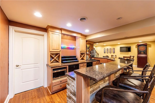 kitchen with light wood finished floors, stainless steel microwave, dark countertops, a kitchen bar, and light brown cabinets
