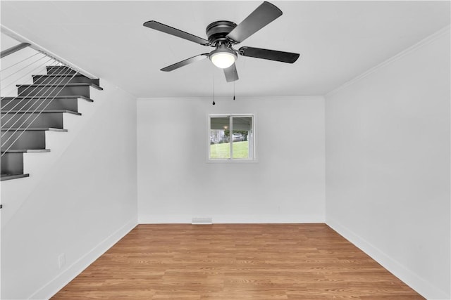 empty room featuring light wood-style flooring, ornamental molding, ceiling fan, baseboards, and stairway