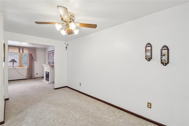 empty room with a fireplace, ceiling fan, light colored carpet, and baseboards