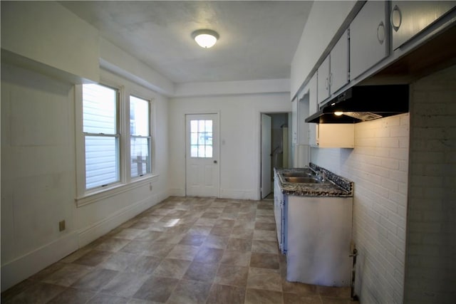 kitchen with dark countertops, a sink, under cabinet range hood, and baseboards