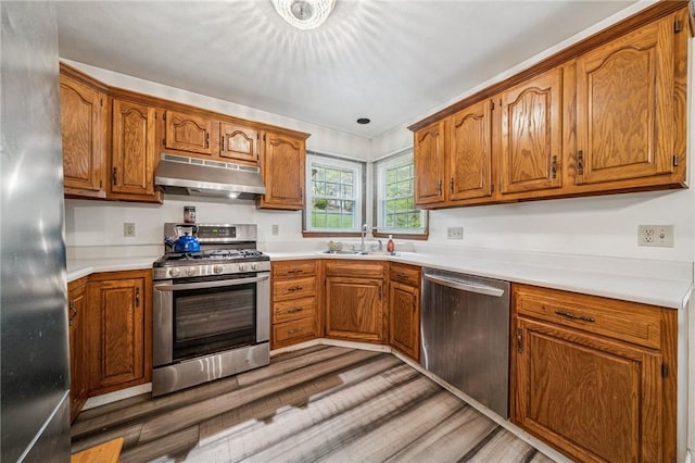 kitchen with brown cabinets, a sink, appliances with stainless steel finishes, under cabinet range hood, and light wood-type flooring