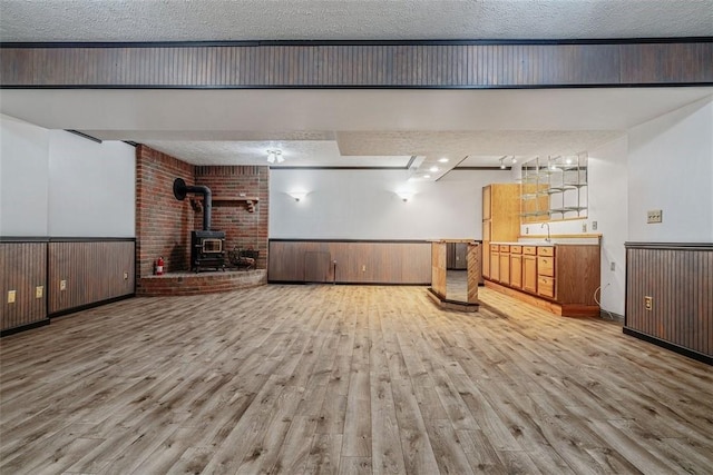 below grade area featuring light wood-type flooring, a wood stove, a textured ceiling, and wainscoting