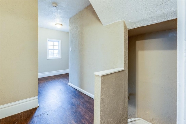 hallway with a textured ceiling, a textured wall, wood finished floors, and baseboards