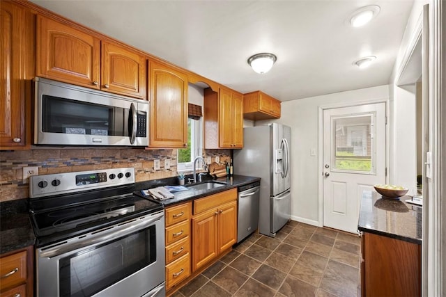 kitchen with a sink, stainless steel appliances, dark stone countertops, brown cabinetry, and backsplash