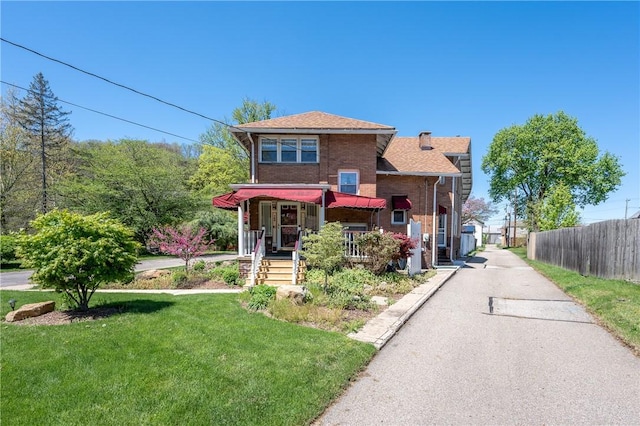 view of front facade with a porch, a front yard, brick siding, fence, and a shingled roof