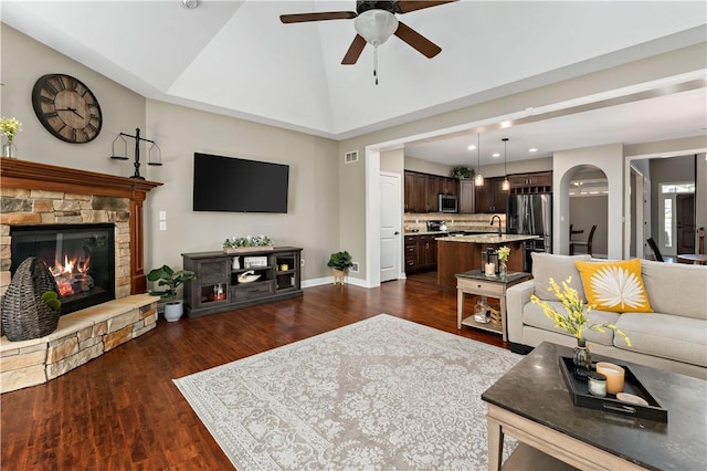 living area with ceiling fan, a stone fireplace, baseboards, dark wood-type flooring, and vaulted ceiling