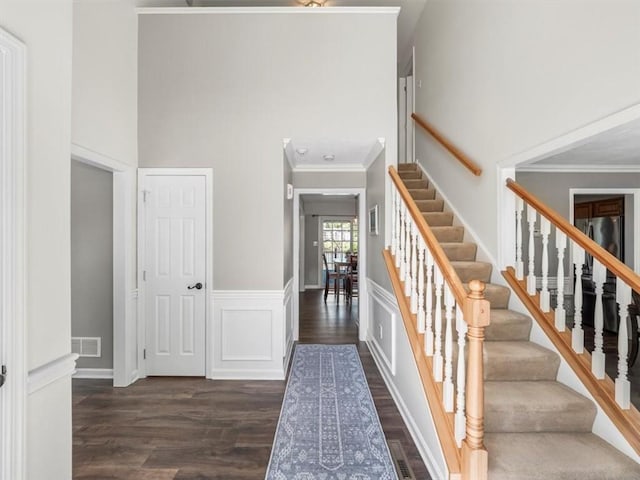 staircase with a high ceiling, wainscoting, wood finished floors, visible vents, and ornamental molding