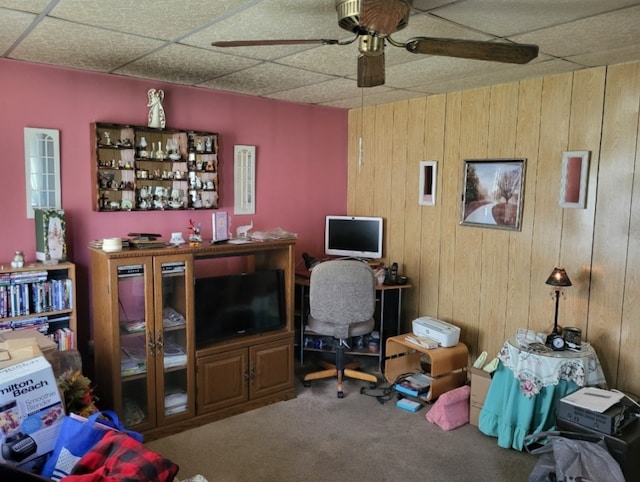 carpeted home office with wood walls, ceiling fan, and a drop ceiling