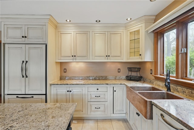 kitchen with cream cabinets, light stone counters, a sink, paneled refrigerator, and glass insert cabinets