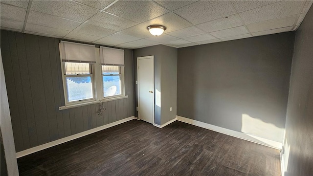 empty room with dark wood-style flooring, baseboards, wood walls, and a drop ceiling
