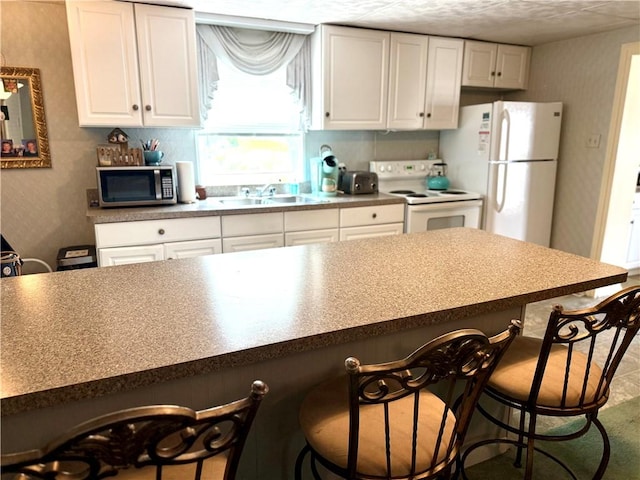 kitchen featuring white cabinets, a sink, white appliances, wallpapered walls, and a kitchen bar