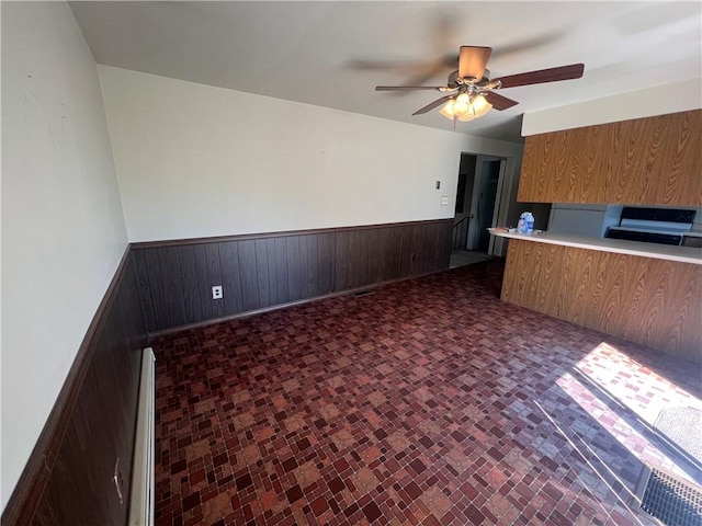 unfurnished living room featuring a ceiling fan, baseboard heating, wood walls, a wainscoted wall, and brick patterned floor