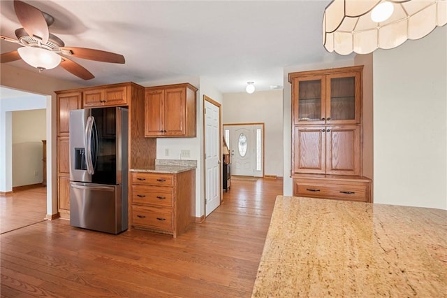 kitchen with ceiling fan, brown cabinetry, light stone countertops, wood finished floors, and stainless steel fridge