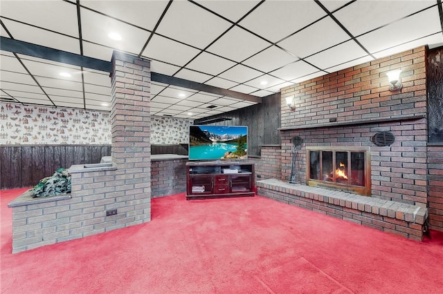 carpeted living room with wooden walls, a brick fireplace, a paneled ceiling, a wainscoted wall, and decorative columns