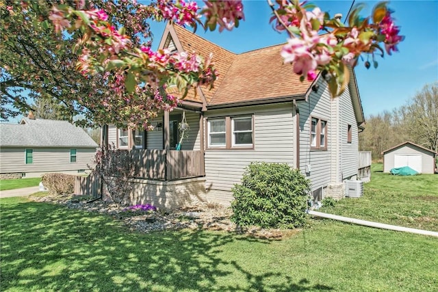 new england style home featuring an outdoor structure, a front yard, a shingled roof, central air condition unit, and a storage shed