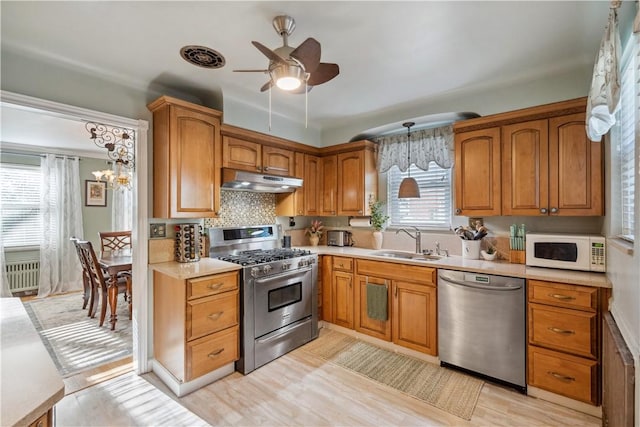 kitchen featuring brown cabinetry, a sink, ceiling fan with notable chandelier, under cabinet range hood, and stainless steel appliances
