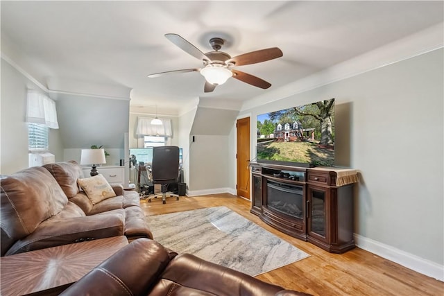 living area featuring ornamental molding, a glass covered fireplace, baseboards, light wood-type flooring, and a ceiling fan