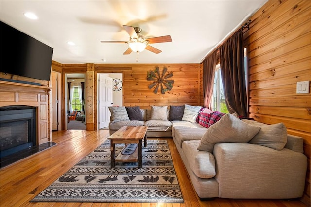 living room featuring wood-type flooring, a glass covered fireplace, ceiling fan, wooden walls, and recessed lighting