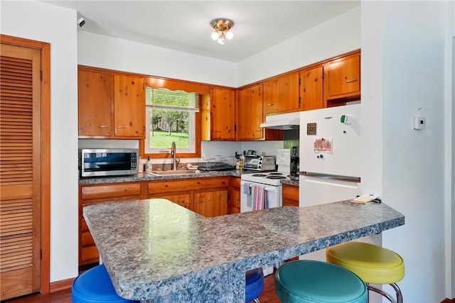 kitchen with a kitchen breakfast bar, brown cabinetry, a sink, under cabinet range hood, and white appliances