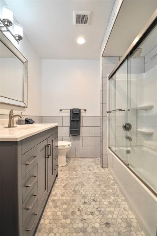 bathroom featuring visible vents, vanity, toilet, tile walls, and combined bath / shower with glass door