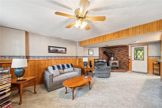carpeted living room featuring wooden walls, a wood stove, a textured ceiling, a wainscoted wall, and a ceiling fan