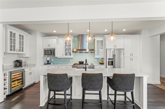 kitchen with appliances with stainless steel finishes, dark wood-style floors, beverage cooler, wall chimney exhaust hood, and an island with sink