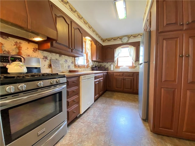 kitchen featuring appliances with stainless steel finishes, a sink, light countertops, under cabinet range hood, and wallpapered walls