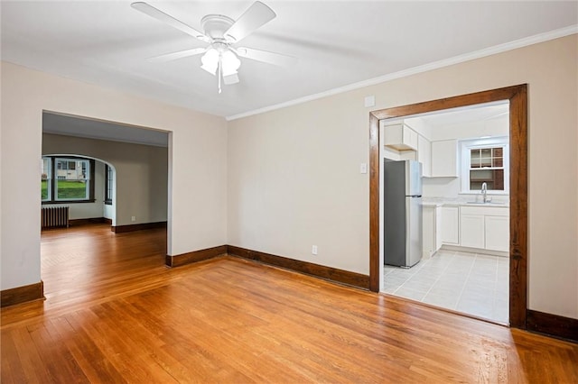 empty room with arched walkways, light wood-type flooring, a sink, baseboards, and radiator heating unit
