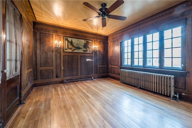 unfurnished room featuring a ceiling fan, radiator heating unit, wooden walls, light wood-type flooring, and a baseboard radiator