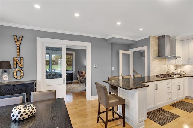 kitchen featuring light wood-style flooring, a peninsula, wall chimney exhaust hood, and white cabinetry